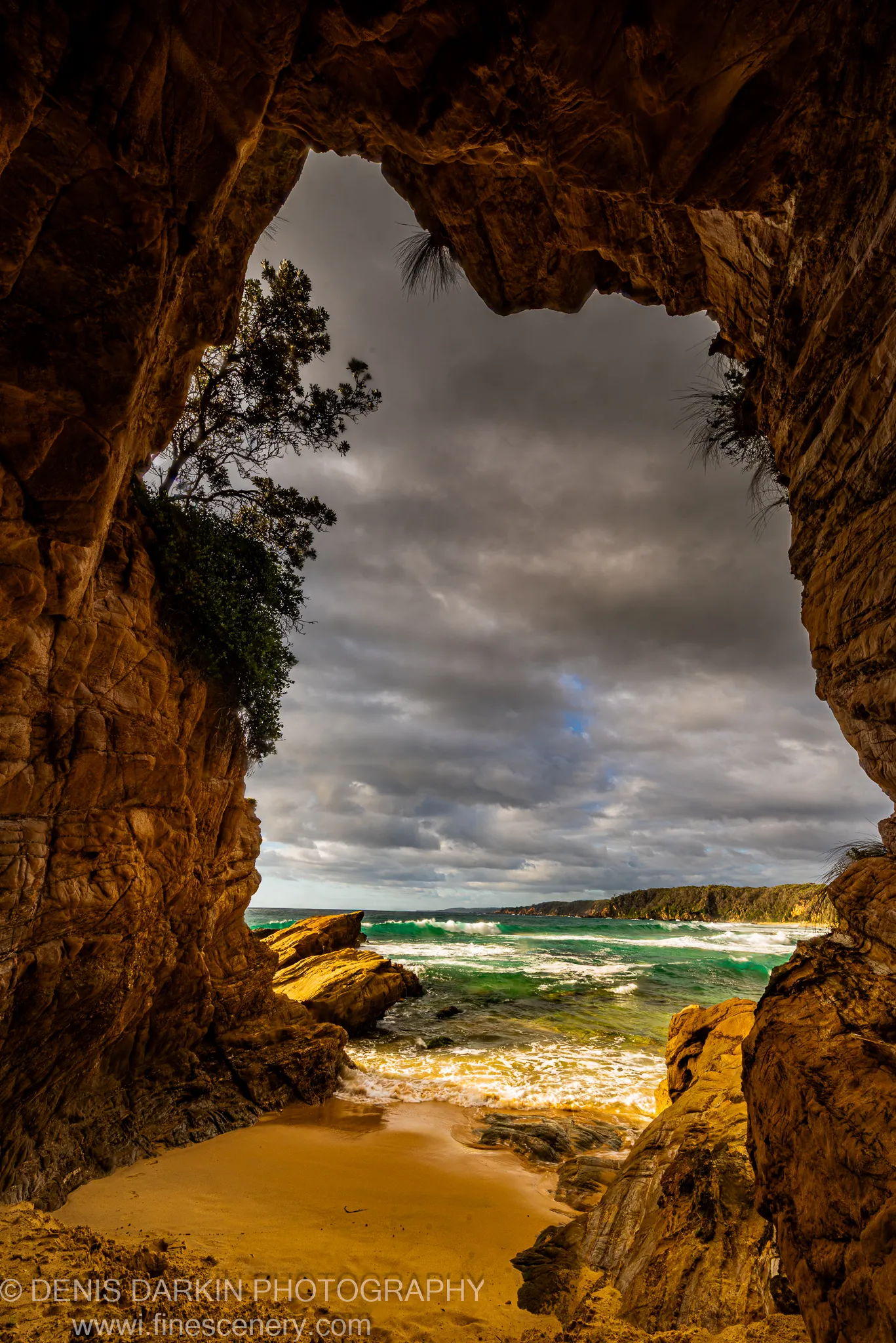 Warm amber light fills a sandstone cave opening to turquoise surf and a sandy beach below storm clouds, South Coast, New South Wales.