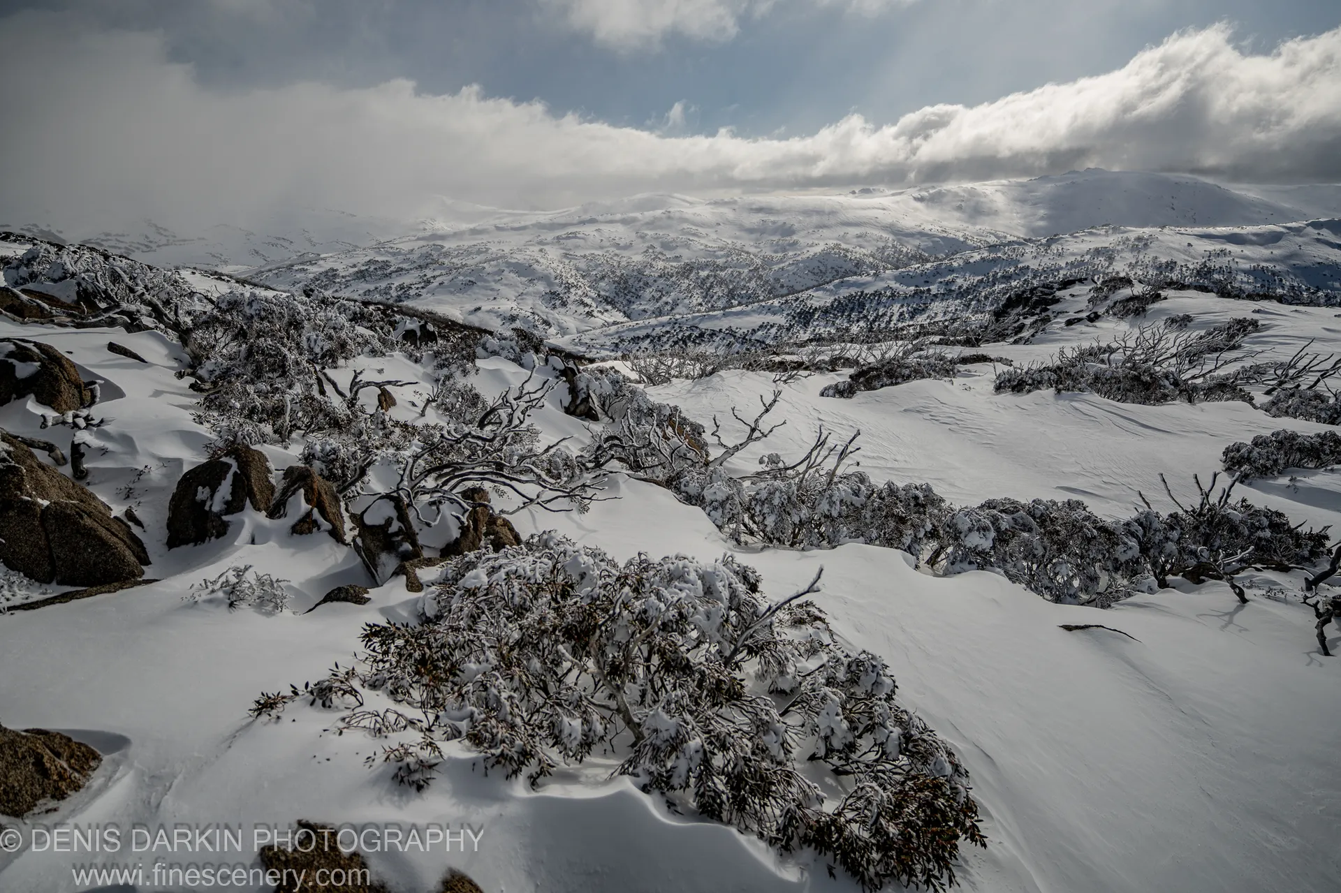 Snow covers dark granite boulders and frost-laden shrubs in the foreground at Perisher Valley, with rolling mountain ridges fading into blue haze.