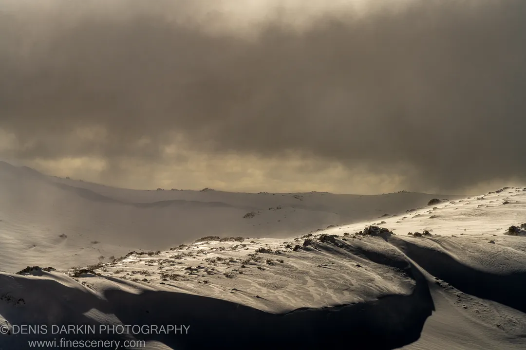 Broken storm clouds part over a rocky alpine ridge at Perisher Valley, sending a shaft of warm light across the wind-scoured snow.