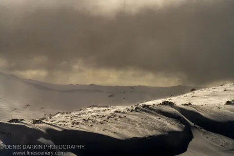 Broken storm clouds part over a rocky alpine ridge at Perisher Valley, sending a shaft of warm light across the wind-scoured snow.