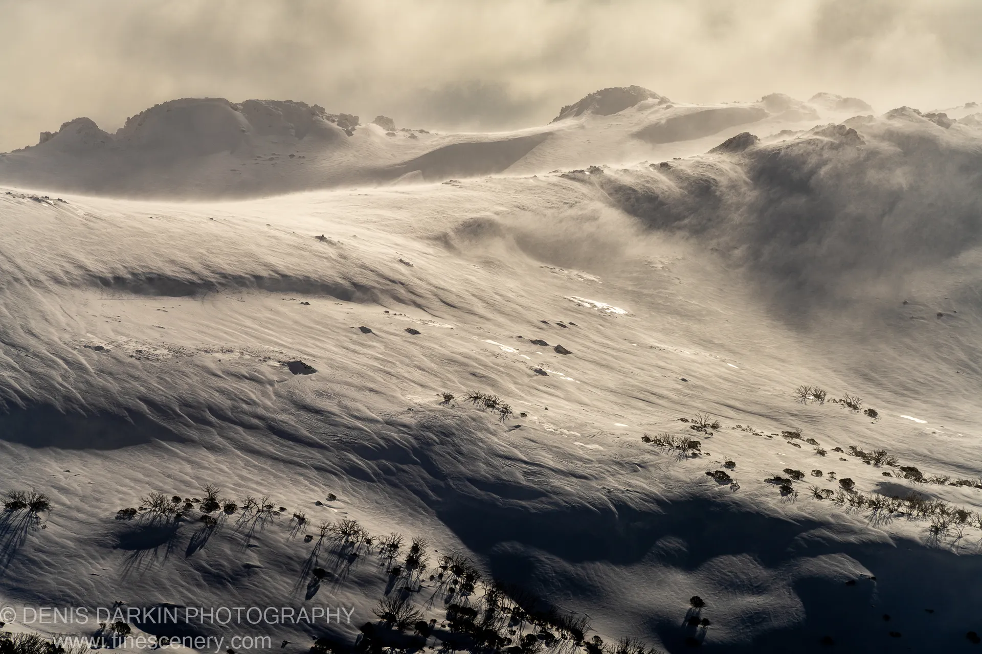 Warm hazy afternoon light falls across snow-covered slopes at Perisher, casting long shadows from sparse vegetation toward distant rocky peaks.