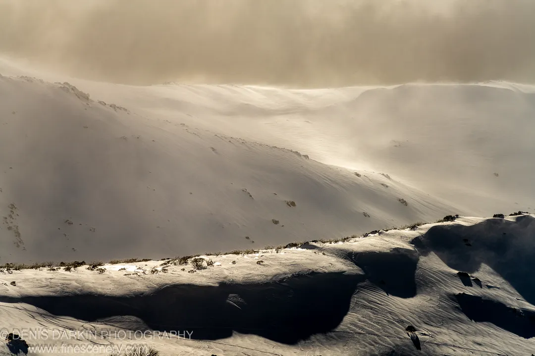 Snow-covered mountain ridgeline at Perisher Valley in winter afternoon light, with long shadows and a warm amber haze above the distant slopes.