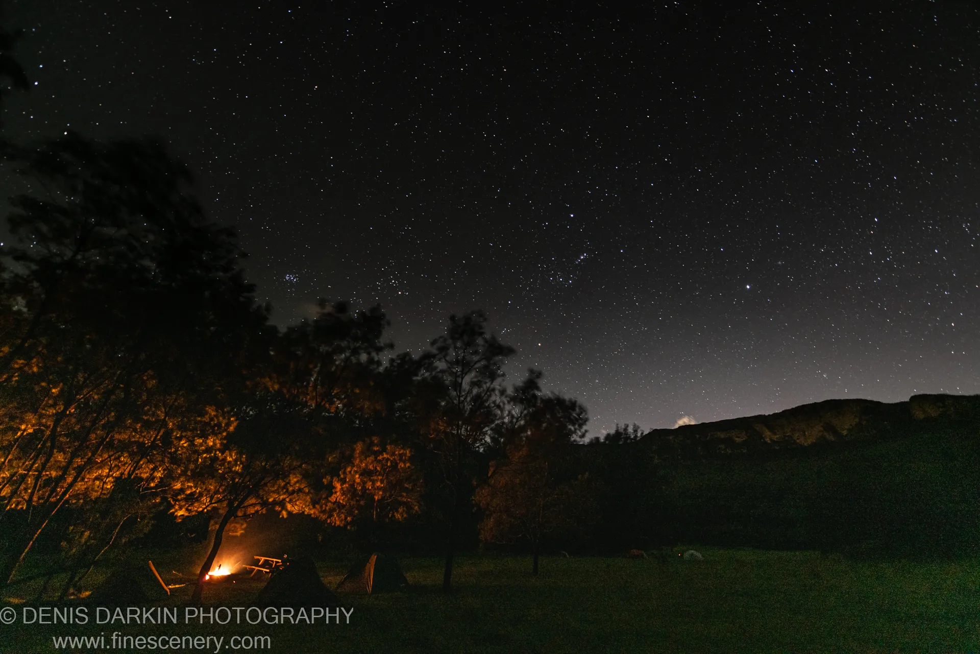 A campfire burns in a grassy clearing in Kedumba Valley, with tents pitched nearby and the Milky Way arching overhead on a spring night.