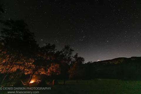 A campfire burns in a grassy clearing in Kedumba Valley, with tents pitched nearby and the Milky Way arching overhead on a spring night.