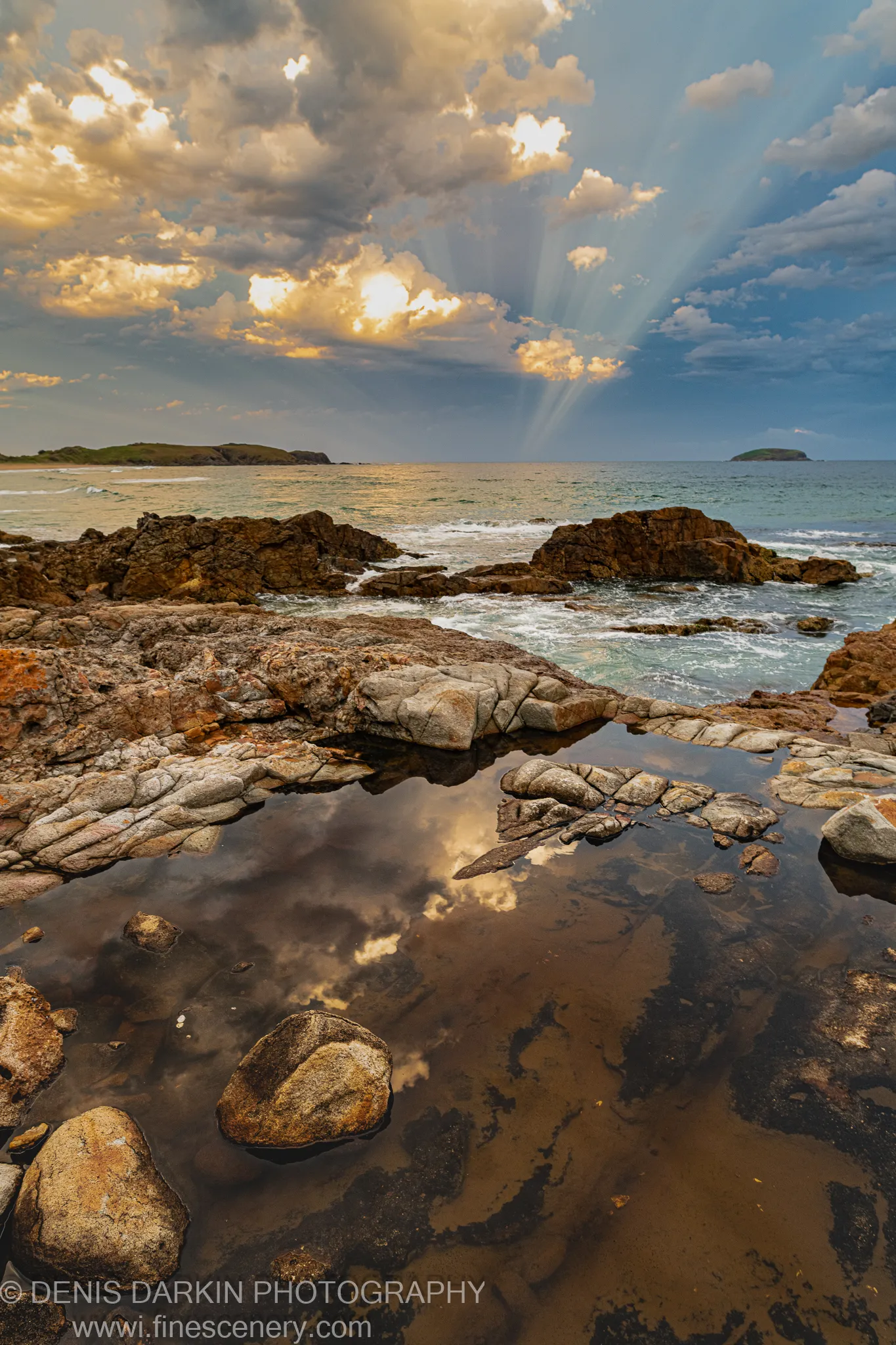 A dark tide pool near Coffs Harbor reflects the warm evening sky, surrounded by rough layered basalt rocks and looking toward distant headlands.