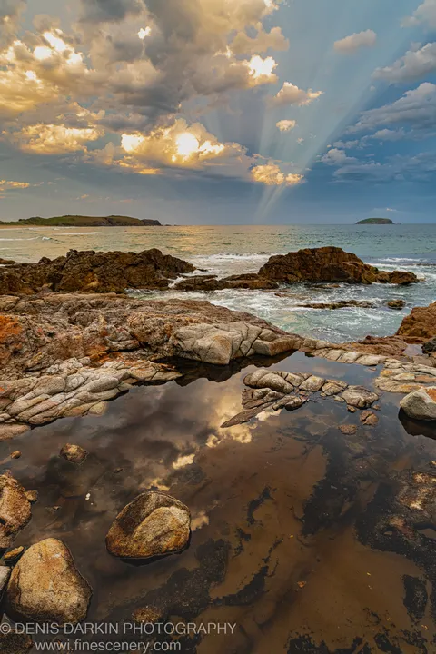 A dark tide pool near Coffs Harbor reflects the warm evening sky, surrounded by rough layered basalt rocks and looking toward distant headlands.