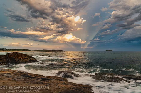 Crepuscular rays cut through storm clouds over the ocean at dusk near Coffs Harbor, with barnacle-covered rocks and wave foam below.