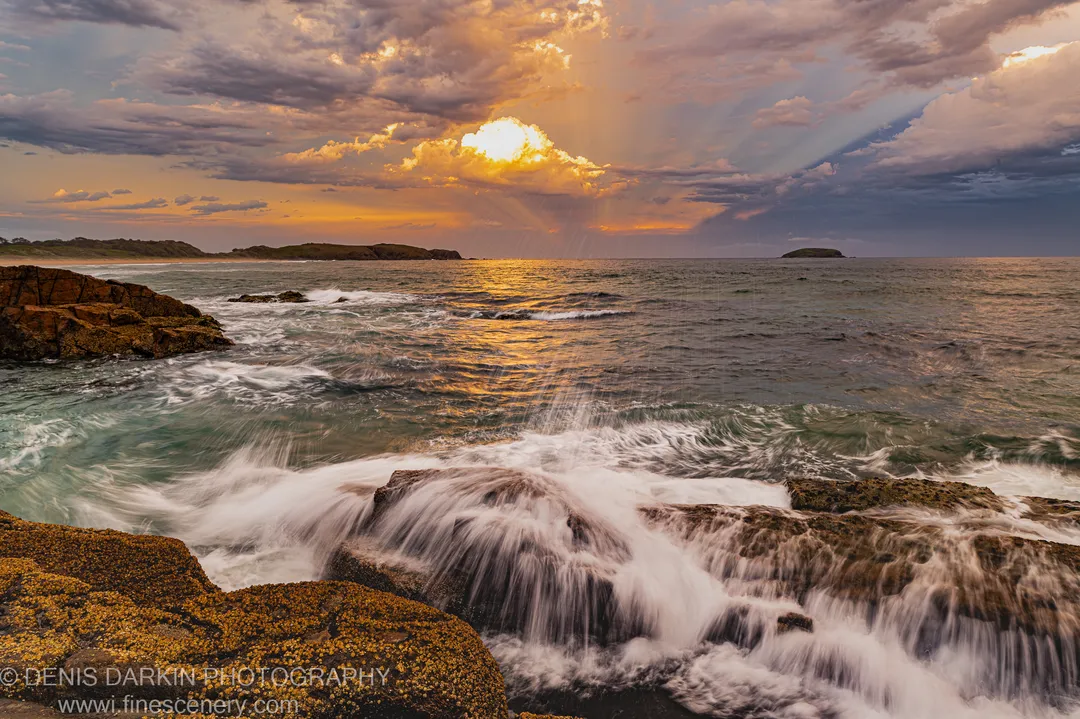 Sunbeams cut through heavy cloud above the ocean at dusk near Coffs Harbor, while smooth wave wash flows across lichen-covered coastal rocks in the foreground.