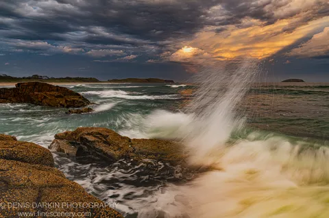 A wave crashes against barnacle-covered rocks at Coffs Harbor, sending spray into a stormy dusk sky with a break of warm light.