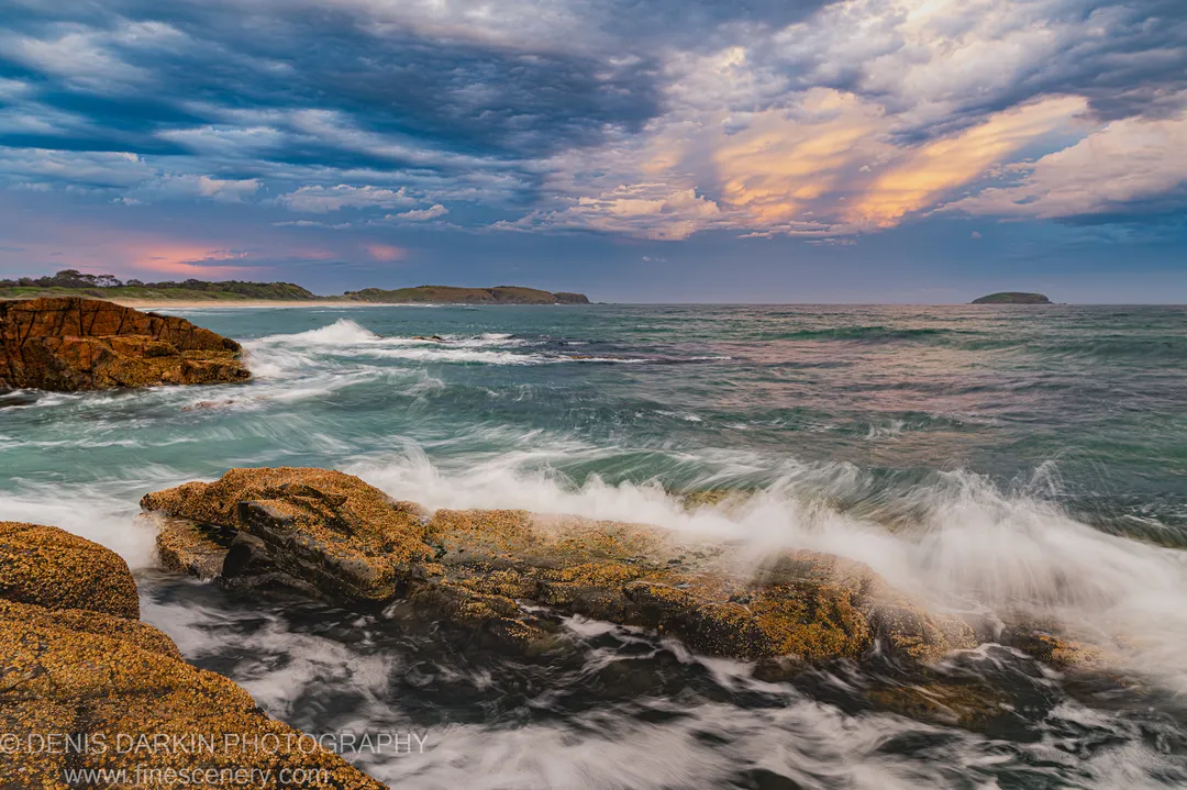 Barnacle-encrusted rocks near Coffs Harbor at dusk, with white water rushing past and storm clouds lit by sunset on the horizon.