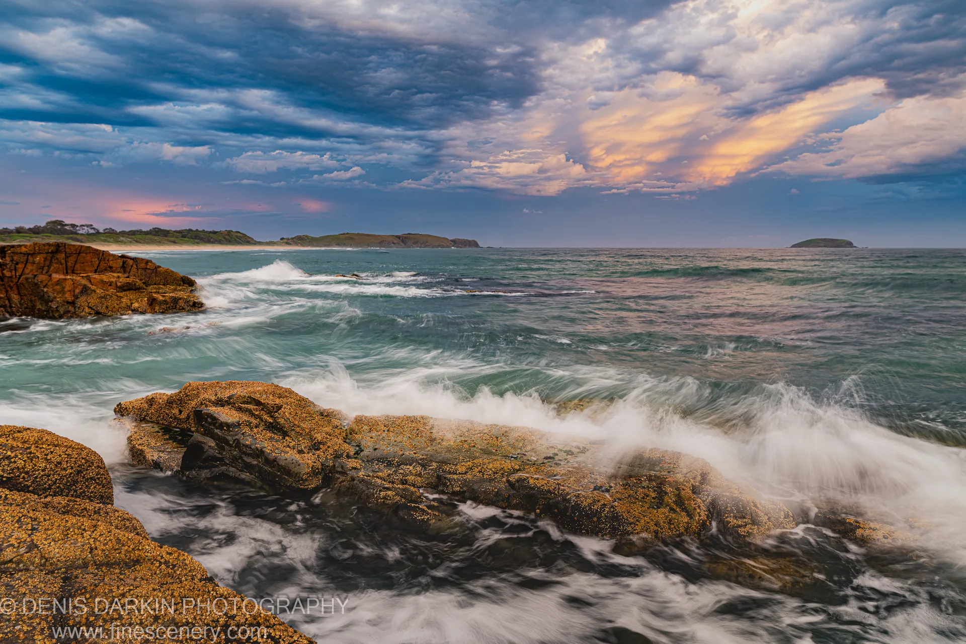 Barnacle-encrusted rocks near Coffs Harbor at dusk, with white water rushing past and storm clouds lit by sunset on the horizon.
