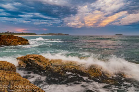 Barnacle-encrusted rocks near Coffs Harbor at dusk, with white water rushing past and storm clouds lit by sunset on the horizon.