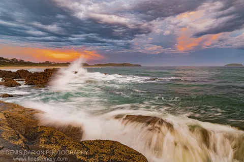 Dark storm clouds over the Coffs Harbor coast at dusk, with crashing waves on lichen-covered rocks and orange sunset light at the horizon.