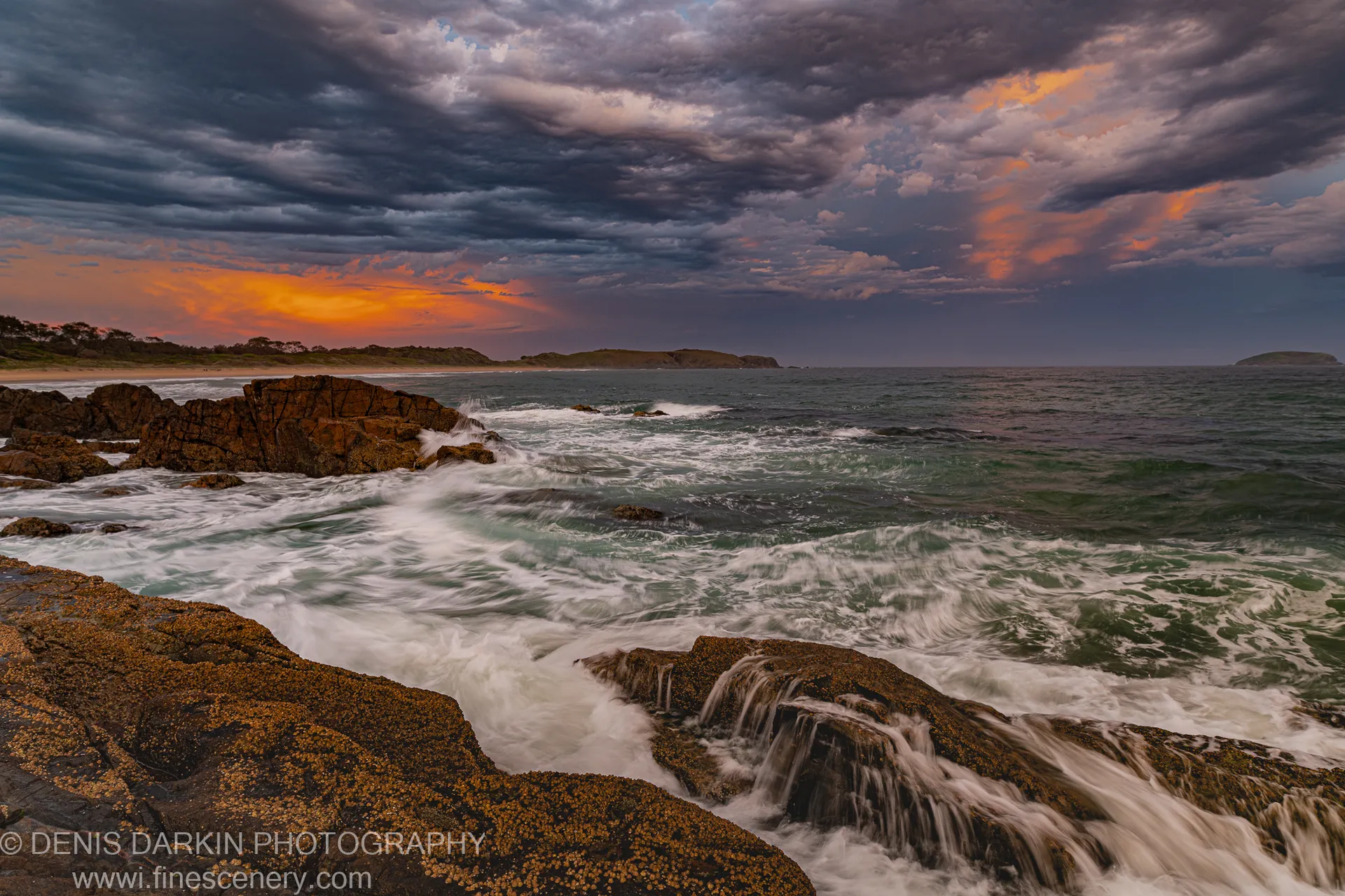 White sea foam swirls around dark barnacle-covered rocks at Coffs Harbor at dusk, with storm clouds split by a low orange sunset on the horizon.