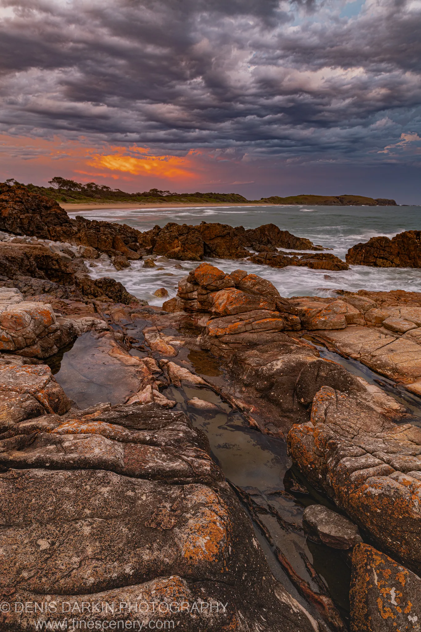 Lichen-streaked rock formations and coastal pools at dusk on the Coffs Harbor shoreline, orange sunset breaking through heavy dark storm clouds.