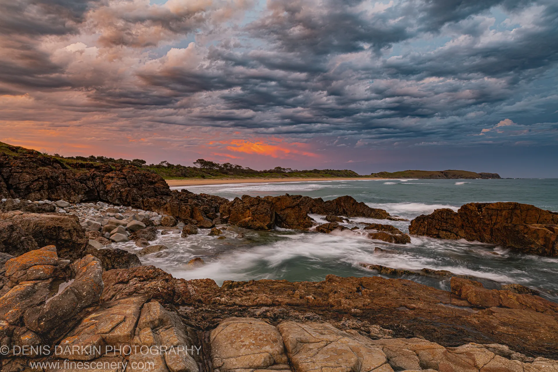 Lichen-speckled rock formations at the edge of a curved bay under dark storm clouds, orange sunset band at the horizon, Coffs Harbor.