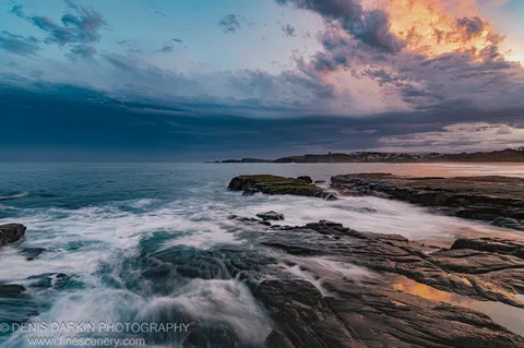 Layered rock formations at blue hour, Coffs Harbor, with deep storm clouds above and warm sunset glow on the horizon.
