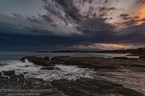 Flat layered rock platforms at dusk, Coffs Harbor, with calm sea and a warm sunset band on the horizon below dark storm clouds.