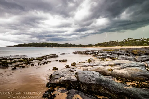Moss-covered tidal rock shelf on the New South Wales coast with two lightning bolts flashing against an afternoon storm sky.