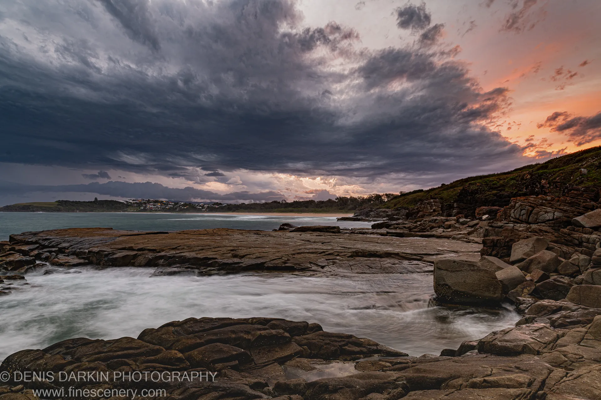 Water flowing over rugged rock at dusk, Coffs Harbor, calm bay behind, dark storm clouds above the warm horizon.