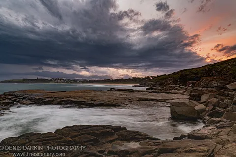 Water flowing over rugged rock at dusk, Coffs Harbor, calm bay behind, dark storm clouds above the warm horizon.