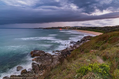 Textured rocks and grassy cliff at dusk, Coffs Harbor, long-exposure ocean smoothed to white, distant town lights along the beach.