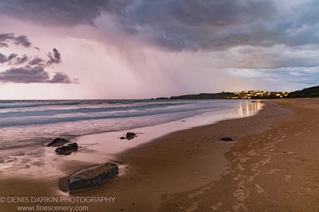 Dark rocks on wet sand at dusk, Coffs Harbor, the sand reflecting warm town lights beneath a purple and blue sky after a rain squall.