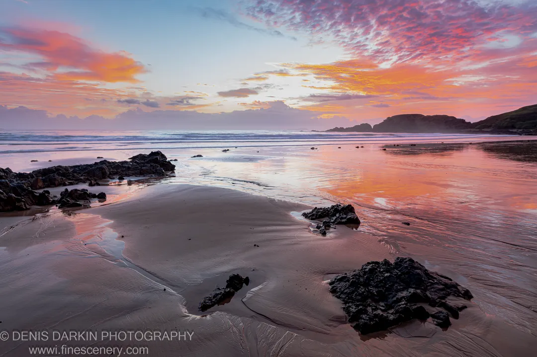 Tidal pools on a rocky Coffs Harbor beach at sunrise, pink and orange sky reflected in shallow water below a silhouetted headland.