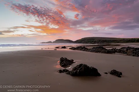 Scattered dark rocks on wet sand near Coffs Harbor at dawn, with pink and orange sunrise sky above distant headlands, New South Wales.