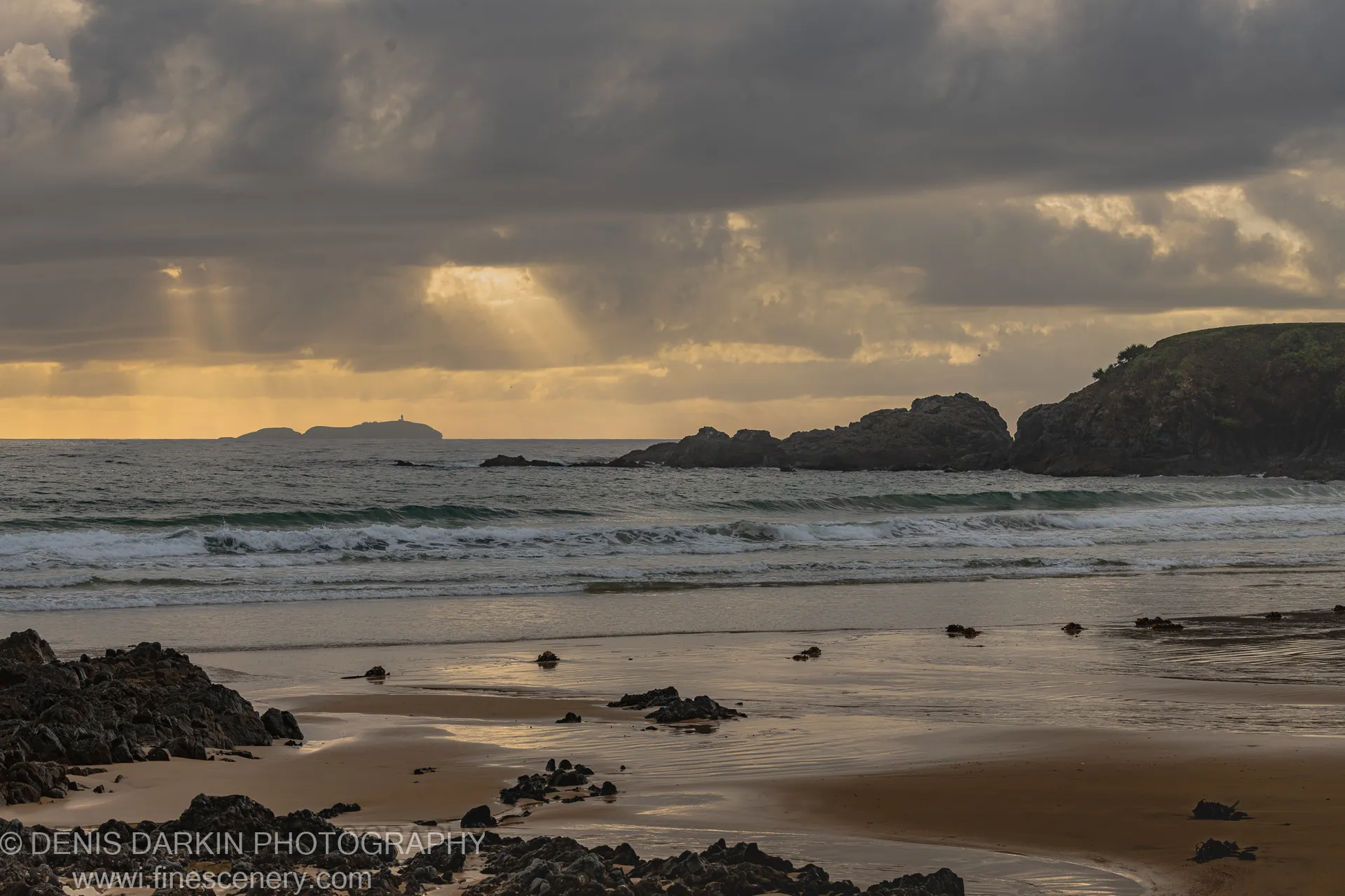 Dark clouds hang over the Coffs Harbor coast as warm light breaks through to the horizon, reflecting in the wet sand below.