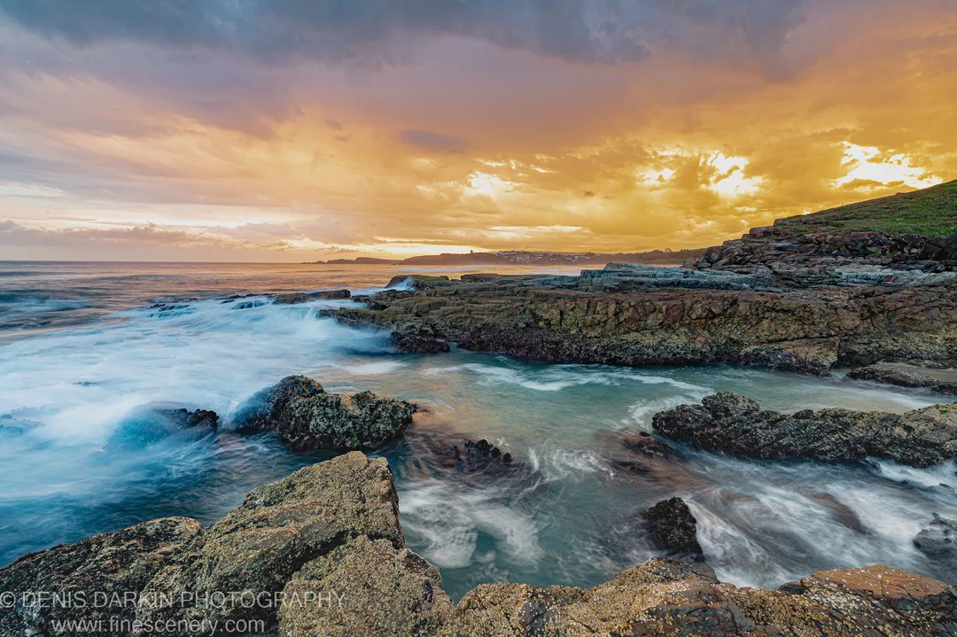 Barnacle-encrusted rock formations at low tide with long-exposure water flowing through tidal channels under a warm golden sky at dusk, Coffs Harbor coast.