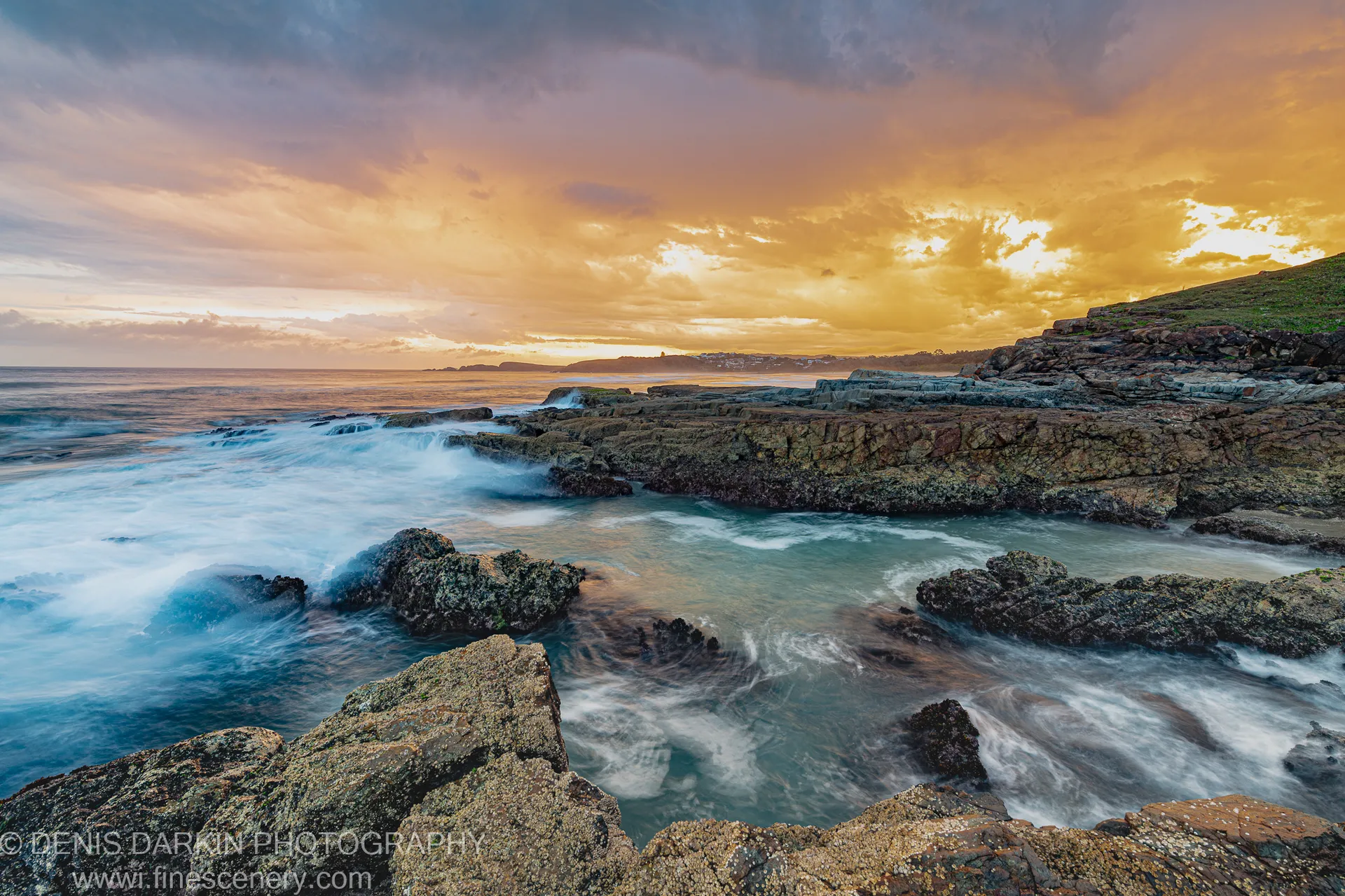 Barnacle-encrusted rock formations at low tide with long-exposure water flowing through tidal channels under a warm golden sky at dusk, Coffs Harbor coast.