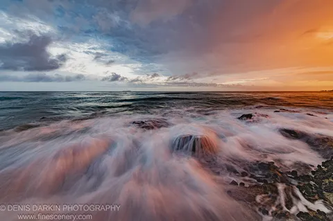 Smooth long-exposure water flows over dark jagged rocks on the Coffs Harbor coast at dusk, under a sky split between blue and warm orange.