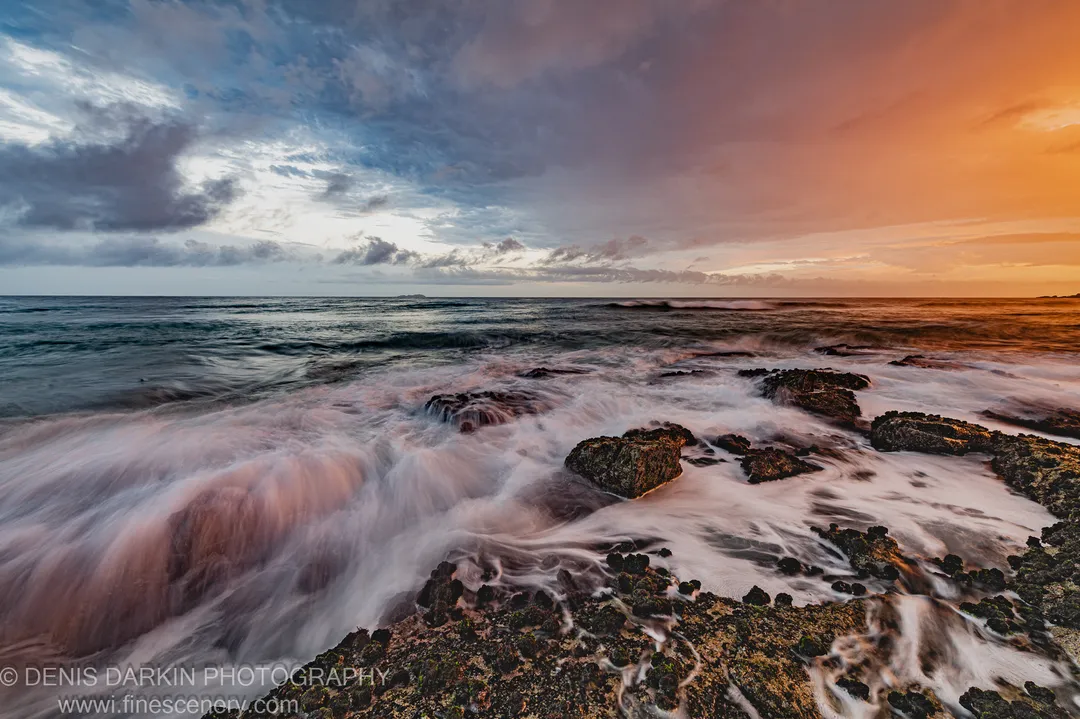 Moss-covered rocks on a coastal shelf with long-exposure surf and a sky shifting from deep grey to burnt orange at the horizon, Coffs Harbor.