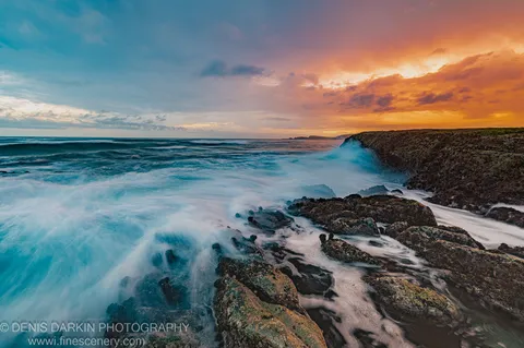 Long-exposure waves wash over moss-covered rocks with a rugged coastal headland visible under a dusk sky ranging from warm orange to cool grey.