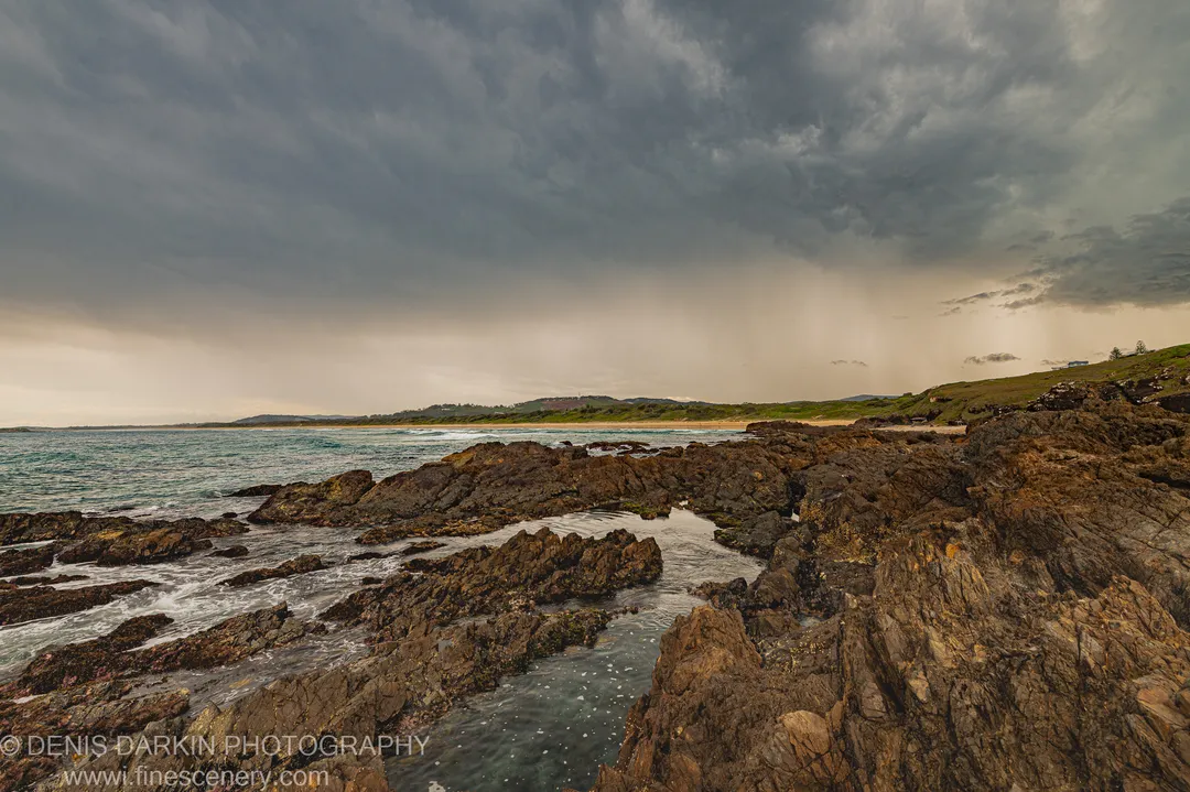 Heavy storm clouds and a curtain of rain hang over dark volcanic rocks and tidal pools at Coffs Harbor, with a sunlit headland beyond.