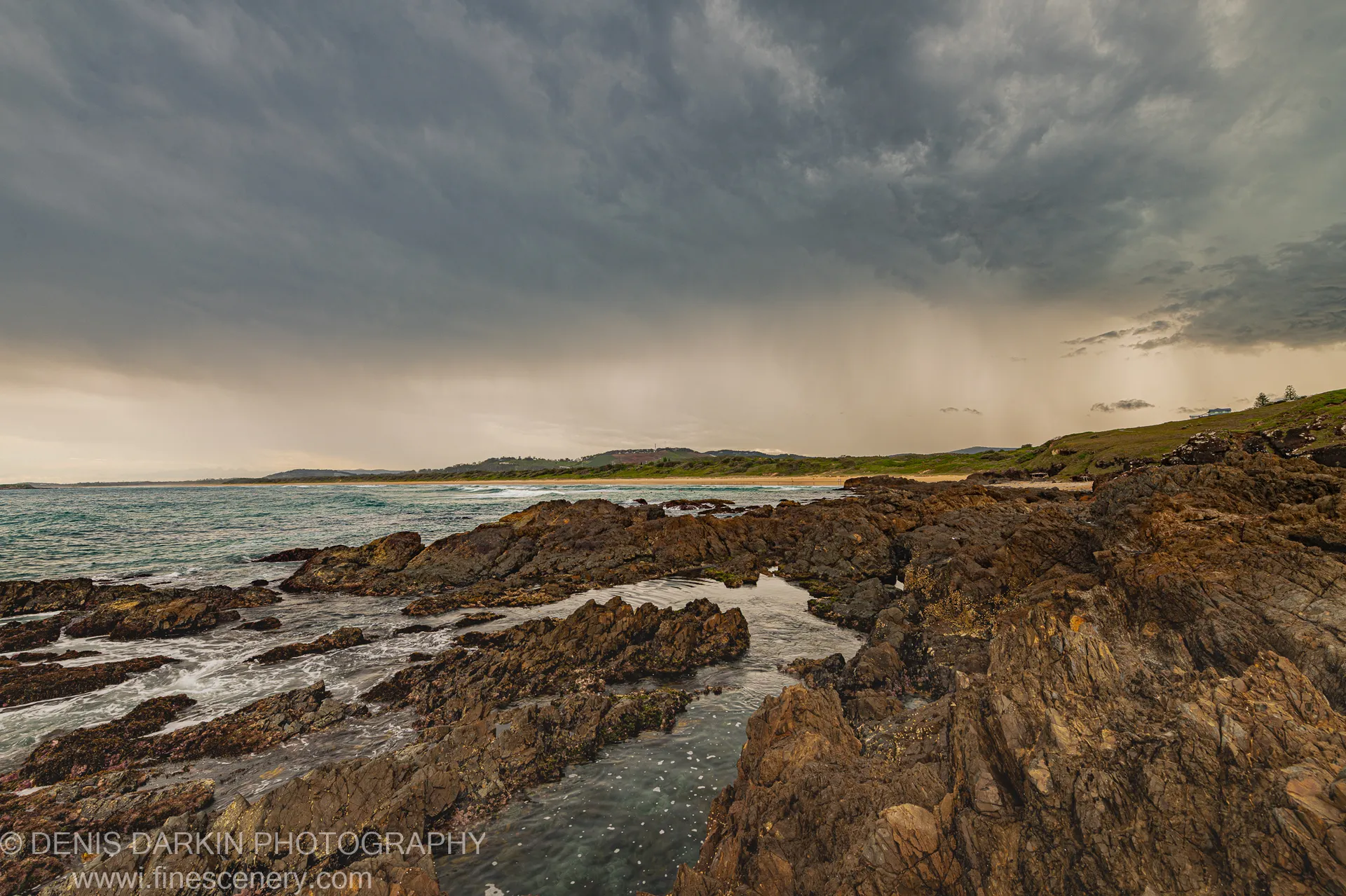 Heavy storm clouds and a curtain of rain hang over dark volcanic rocks and tidal pools at Coffs Harbor, with a sunlit headland beyond.