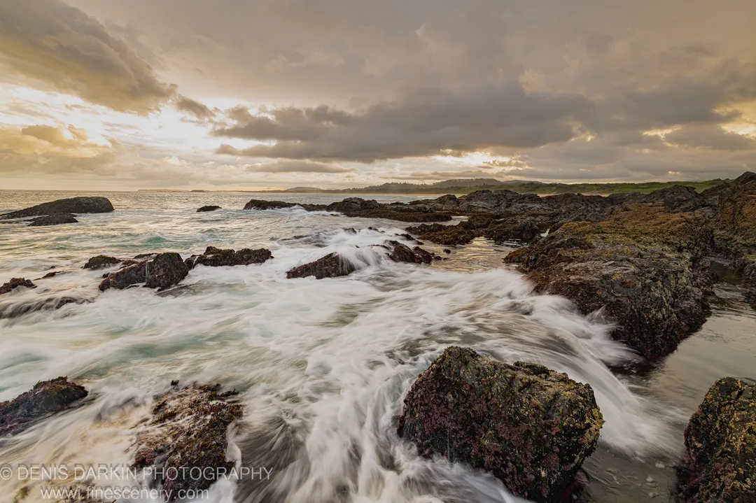 Turbulent water rushes over dark algae-covered boulders along the Coffs Harbor coast, with golden light breaking through the edges of heavy storm clouds above.
