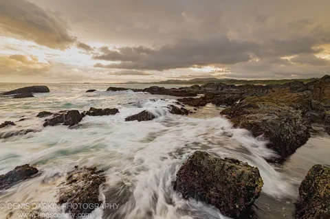Turbulent water rushes over dark algae-covered boulders along the Coffs Harbor coast, with golden light breaking through the edges of heavy storm clouds above.