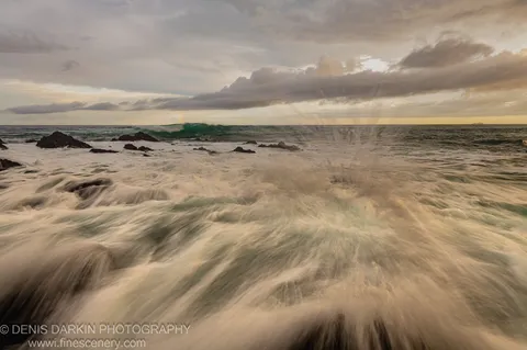 Warm light breaks through a cloud bank at the Coffs Harbor coast, revealing a faint rainbow arc in the mist above dark volcanic rocks.