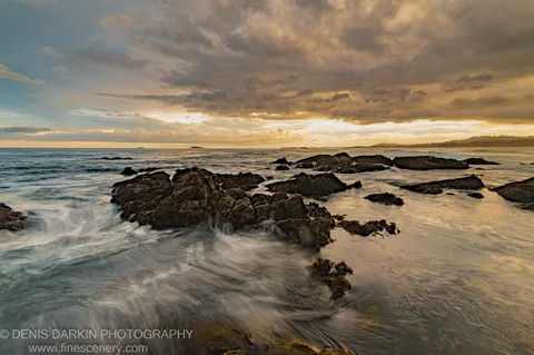 Long-exposure water flows around seaweed-covered rocks at dusk on the Coffs Harbor coast, with deep blue sea and heavy clouds above.