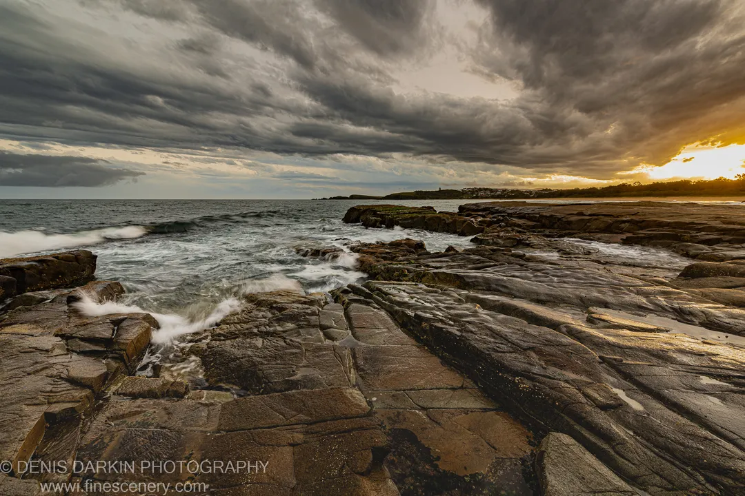 Layered rock in a diagonal plane at the Coffs Harbor coast at dusk, warm light through heavy storm clouds, white foam on wet stone.