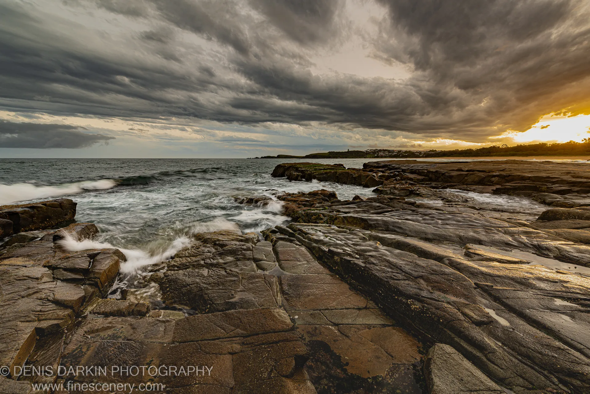 Layered rock in a diagonal plane at the Coffs Harbor coast at dusk, warm light through heavy storm clouds, white foam on wet stone.