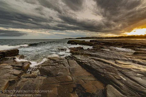 Layered rock in a diagonal plane at the Coffs Harbor coast at dusk, warm light through heavy storm clouds, white foam on wet stone.