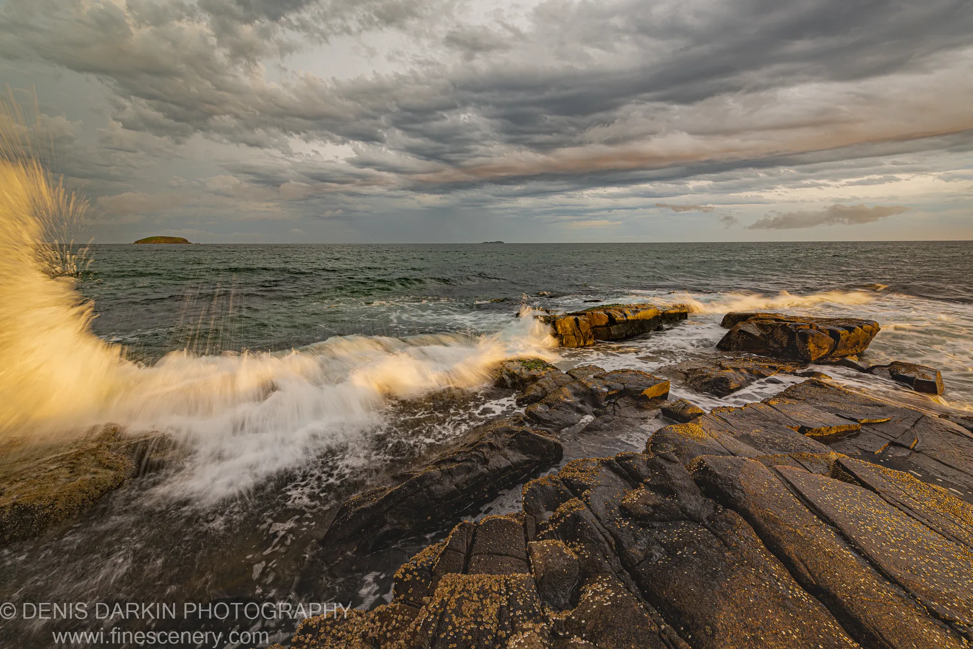 Barnacle-covered layered rock platform at Coffs Harbor dusk, with wave spray and small islands visible under storm clouds.