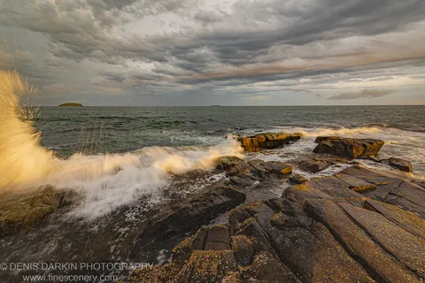 Barnacle-covered layered rock platform at Coffs Harbor dusk, with wave spray and small islands visible under storm clouds.