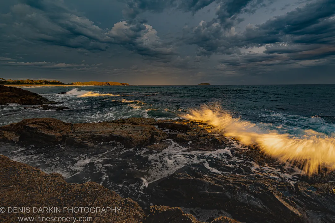 Waves crashing against a rocky Coffs Harbor shoreline at dusk, with golden-lit spray below dark storm clouds and a distant headland.