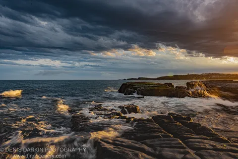 Mossy coastal rocks at Coffs Harbor dusk, with waves below dark storm clouds and warm light breaking on the horizon.