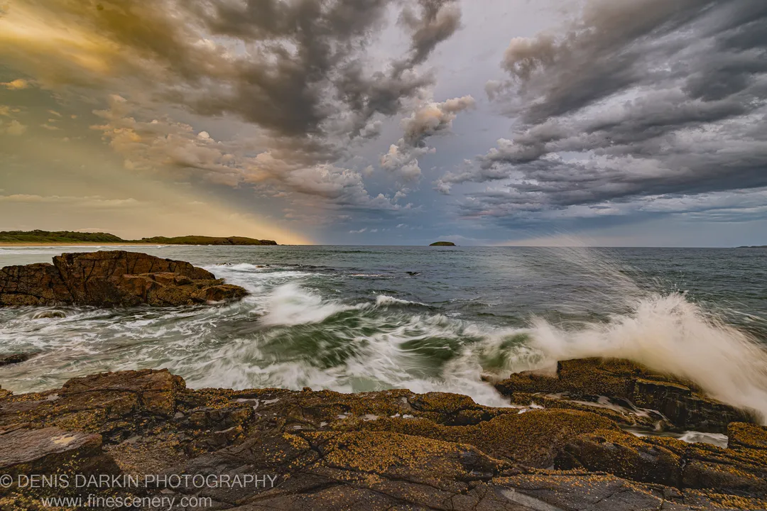 Flat, barnacle-covered rock shelves at Coffs Harbor with wave spray and blurred waves below storm clouds with golden light breaking through, New South Wales.