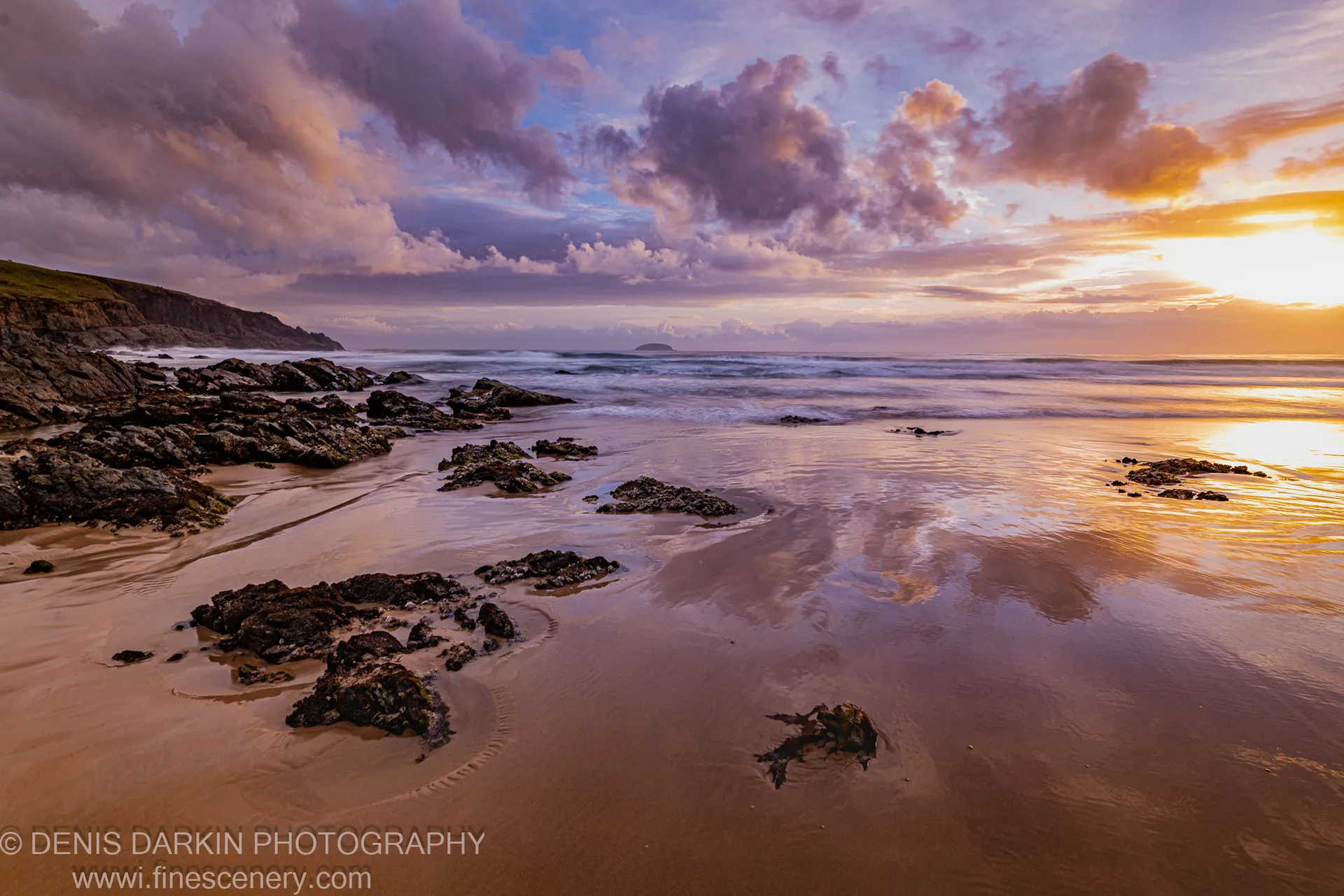 Warm dawn light reflects in wet sand on the Coffs Harbor coast beneath purple clouds and a distant island silhouette on the horizon.